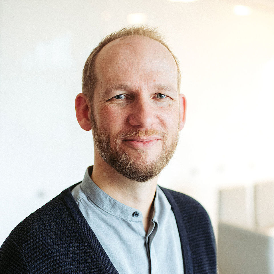 MItarbeiter ambulanter Pflegedienst, kurzes blondes Haar, kurzer Vollbart, hellblaues Hemd, dunkelblaue Strickjaccke, kurzer Bart, freundlich lächenlnd